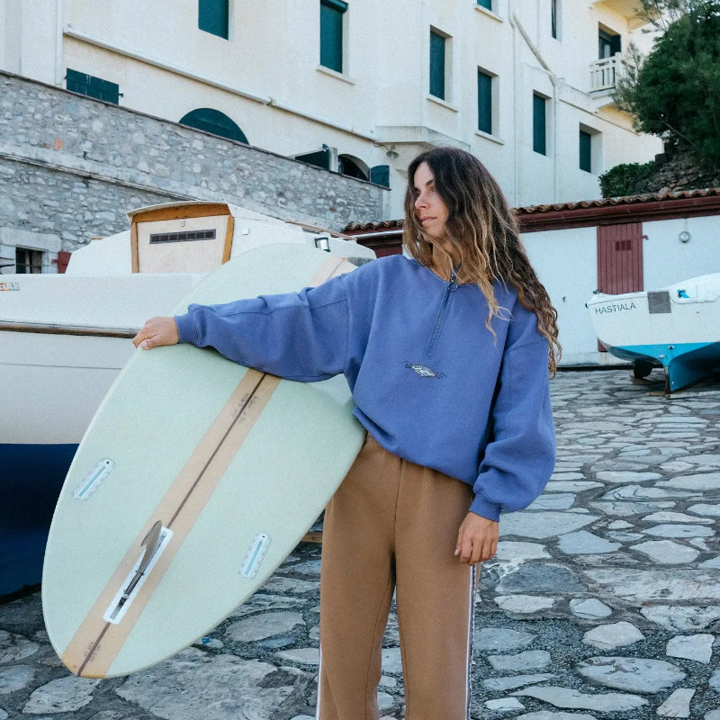 Woman holding a surfboard on a stone street with buildings and boats in the background